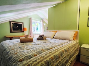 A bedroom with a bed, pillows, a bedside table, a lamp, a framed picture, and a window with curtains.