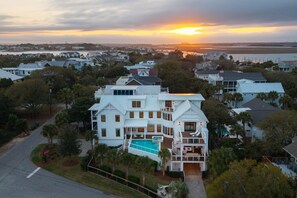 A large white house with a swimming pool, surrounded by trees and a view of the sunset over water. - The Ivory Tide by Charleston Vacays (Isle of Palms)