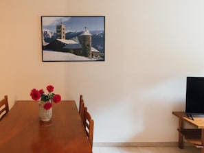 Taula de menjador amb cadires, un vas amb flors vermelles, un quadre a la paret i un televisor sobre un suport.