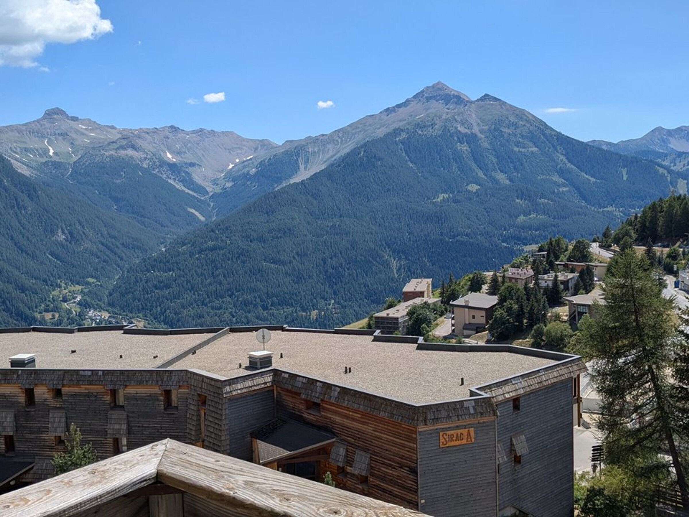 A mountain village with a prominent peak in the background, a wooden building with a sign 'SIRAC A' on the side, and a clear sky.