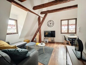 Ett rum i loftstil med synliga träbjälkar, en tv, ett matbord med stolar, en soffa och ett litet runt bord med en lampa.