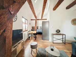 Ett modernt vardagsrum med synliga träbjälkar, en matplats, ett kök och ett litet bord med en dekorativ detalj.