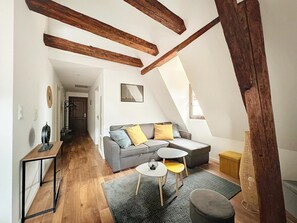 Une pièce de style loft avec des poutres en bois apparentes, un canapé gris agrémenté de coussins jaunes, une petite table ronde et un tapis gris.