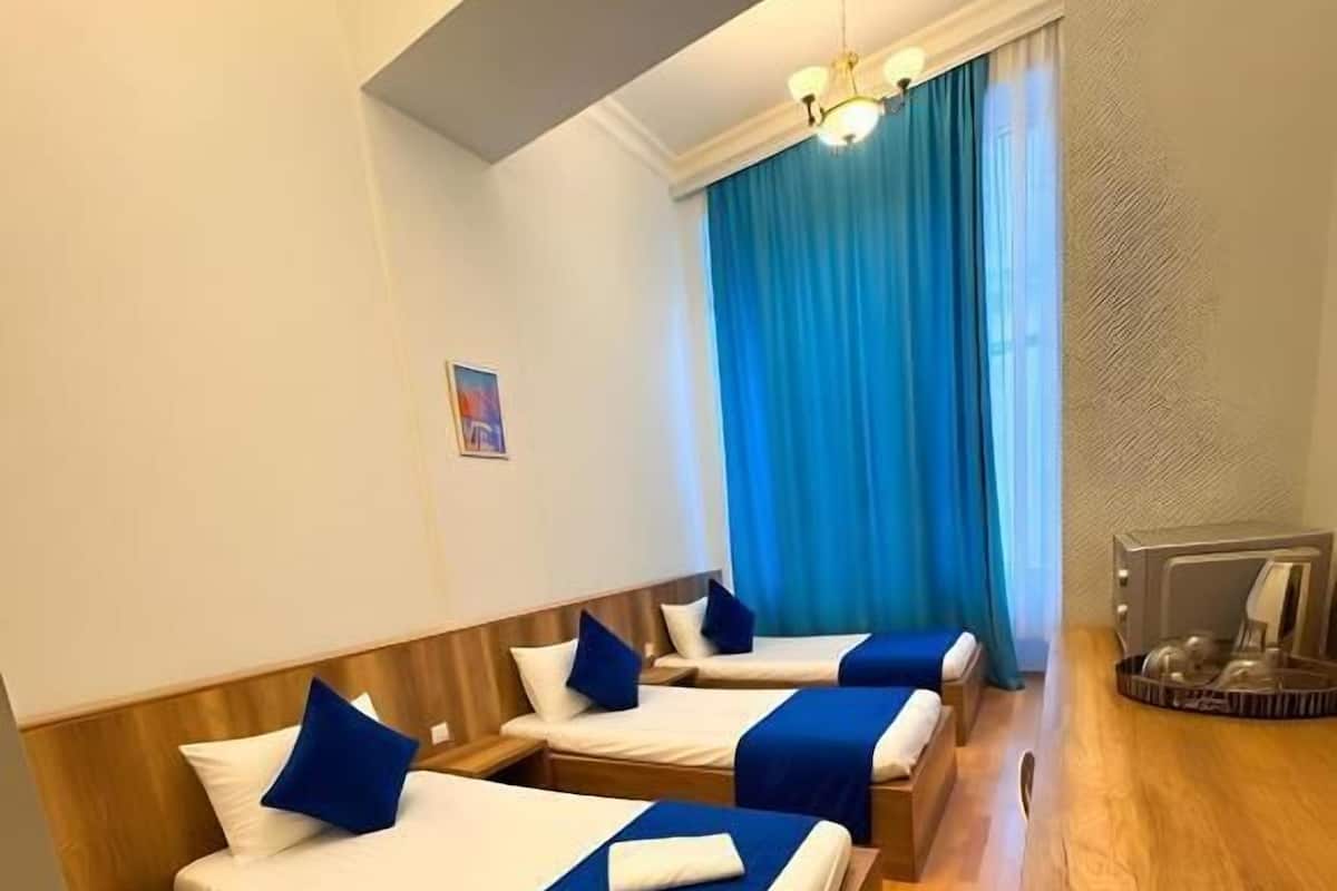 Habitació d'hotel amb tres llits, sòls de fusta, una cortina blava i una taula petita amb una làmpada.