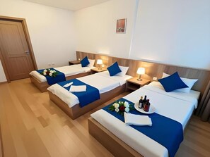 A hotel room with three beds, each with a blue bedspread and white linens, a bedside table with a lamp, and a small table with a flower arrangement and bottles. - Liman Hotel (Baku)
