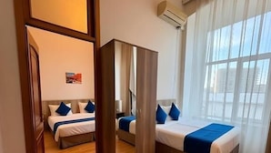 Ett hotellrum med trägolv, en säng med blått sänglinne, en stor spegel och ett fönster med tunna, genomskinliga gardiner.