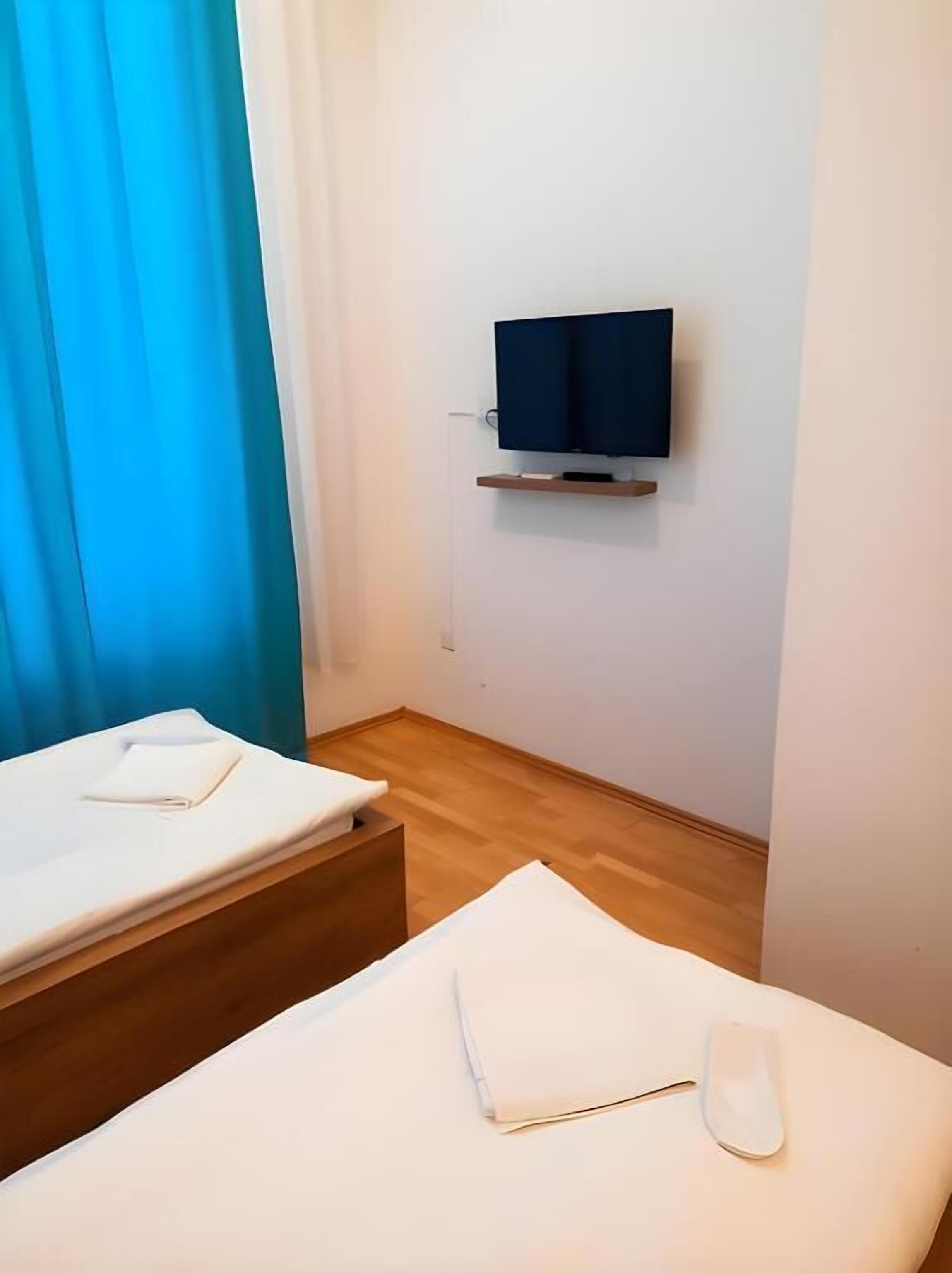A hotel room with a bed, a bedside table, a TV mounted on the wall, and a window with blue curtains.