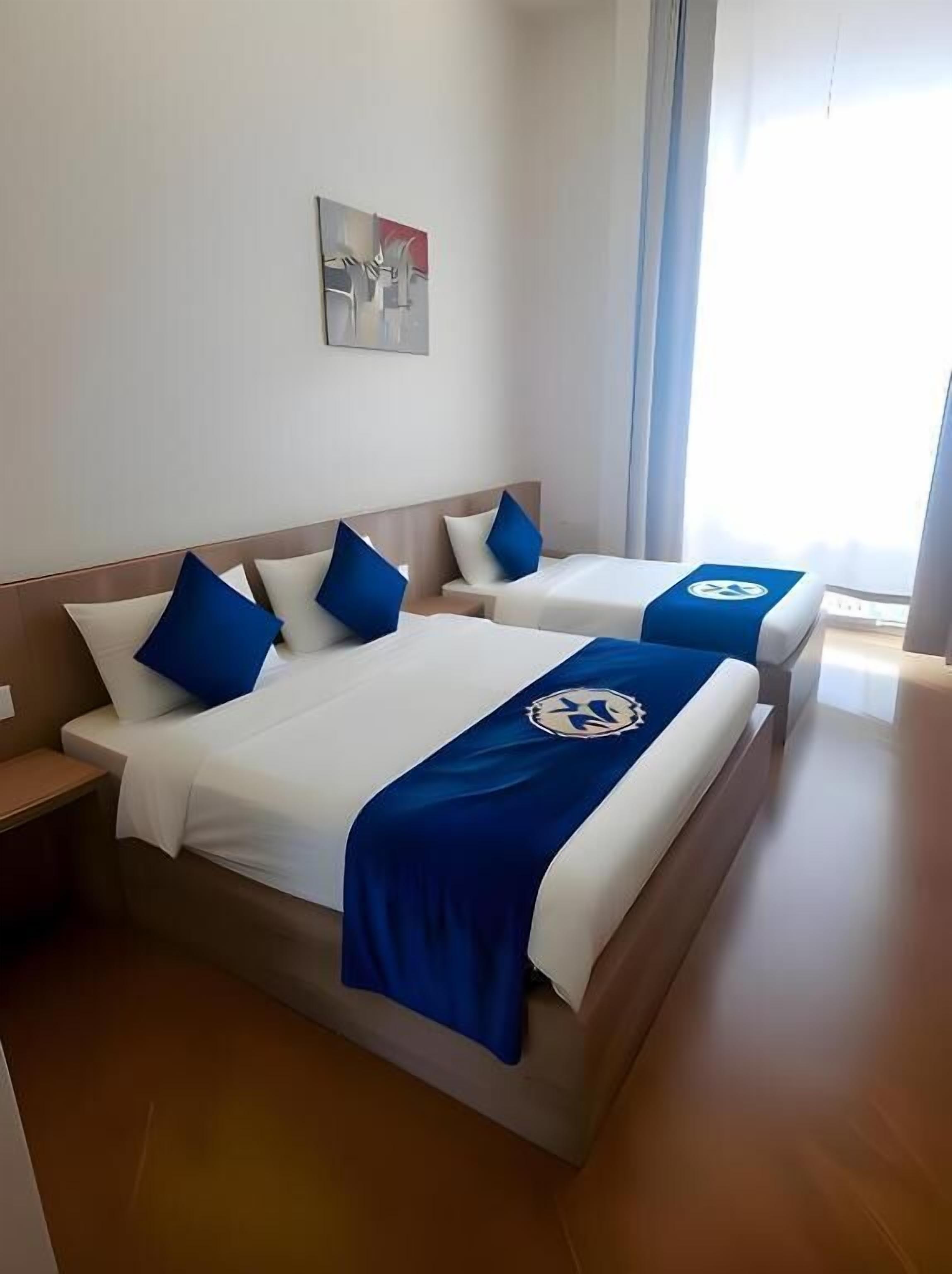 Two beds with blue and white bedding, a small painting on the wall, and a window with curtains.