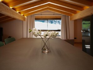 Een vergaderruimte met een lange tafel, een vaas met witte bloemen, houten balken in het plafond en een raam met gordijnen.