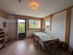 Ein Zimmer mit Tisch, Stühlen, Heizkörper, einem fenster mit Vorhängen und Blick auf Grün.