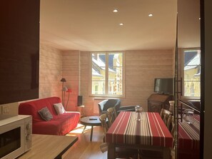 A compact living space with a red sofa, a dining table, a TV, and a window with a view of buildings. - Cozy apartment in the heart of Cauterets - 4 guests (Cauterets)
