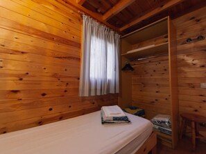 A wooden cabin room with a bed, a shelf with items, and a window with curtains.