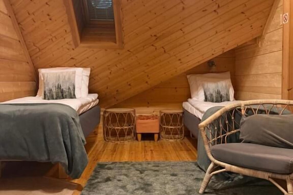 A small, cozy attic bedroom with two beds, a wicker chair, and a small table.