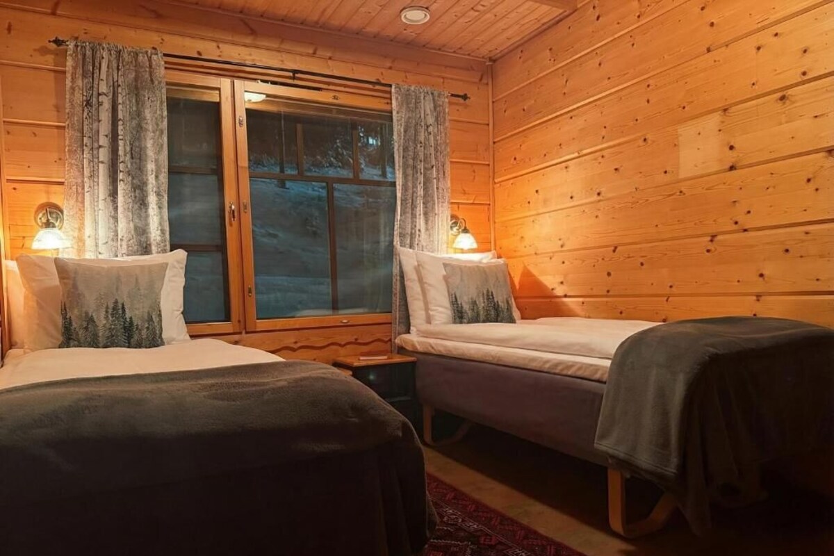 A wooden cabin room with two beds, a window with curtains, and a small table with a lamp.