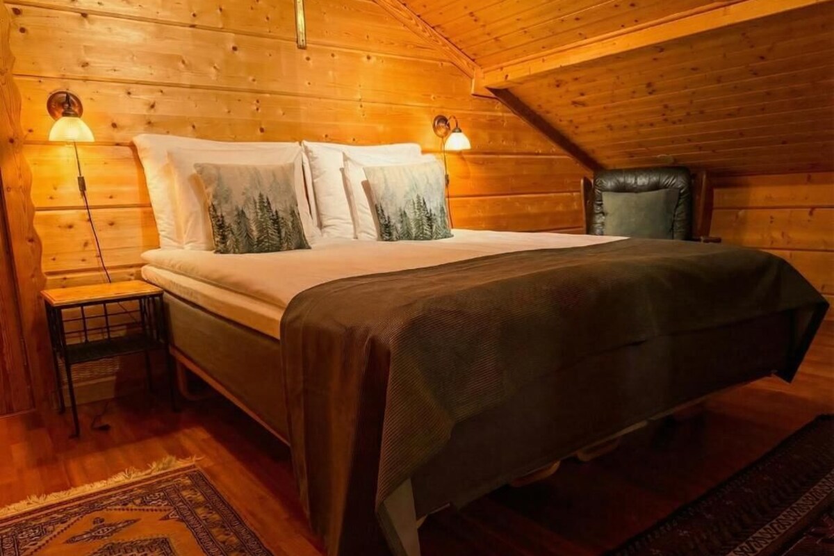 A wooden cabin bedroom with a bed, two wall-mounted lamps, a nightstand, and a patterned rug.