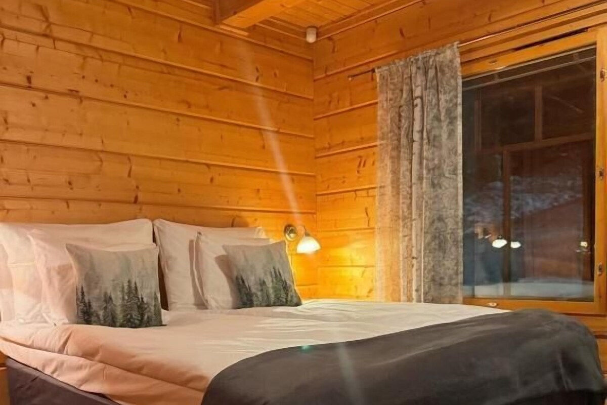 A wooden cabin bedroom with a bed, pillows, a nightstand, and a window with curtains.