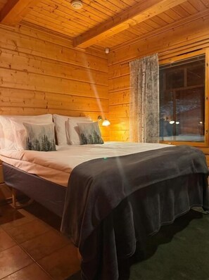 Un dormitorio de cabaña de madera con cama, almohadas, mesita de noche y ventana con cortinas.