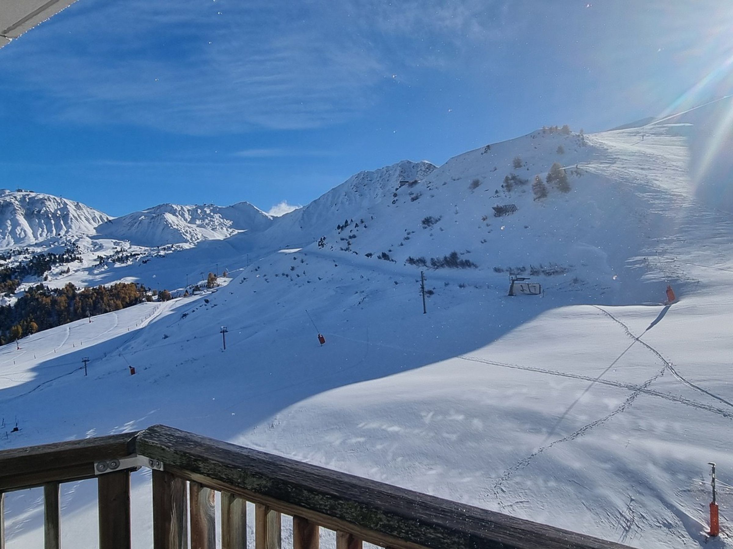 A snowy mountain landscape with ski slopes, a ski lift, and a wooden railing in the foreground.