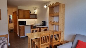 Kompakti keittiö, jossa on puukalusteet, ruokapöytä tuoleineen, sisäänrakennettu hylly ja harmaa sohva, jonka päällä on punainen koristetyyny.
