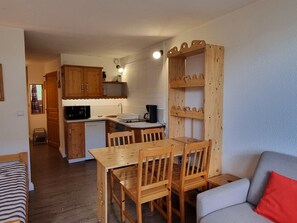 Eine kleine Küche mit Holzausstattung, einem Esstisch mit Stühlen, einer eingebauten Ablage und einem grauen Sofa mit rotem Kissen.