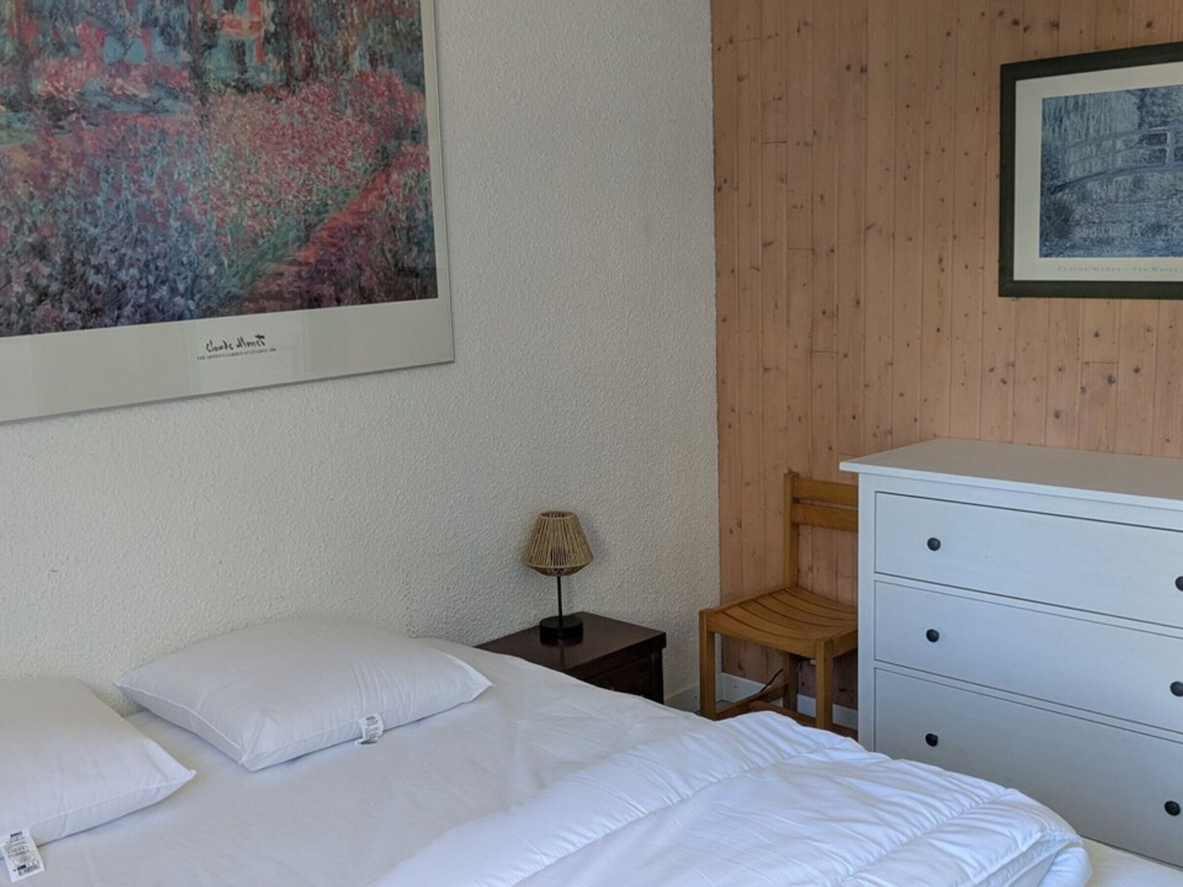 A bedroom with a bed, a nightstand, a lamp, a chair, a dresser, and two framed artworks on the wall.