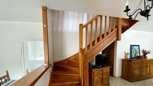 Una escalera de madera baja a un nivel inferior, un comedor con una mesa y sillas, y un mueble con un televisor.