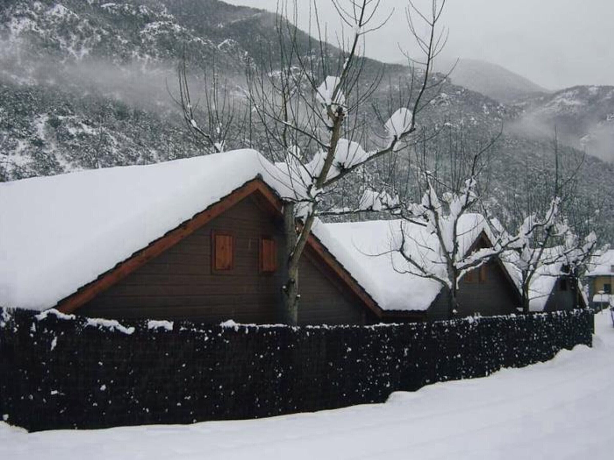 A wooden cabin with a sloped roof, surrounded by snow-covered trees and mountains.