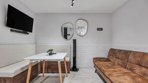 Ein modernes Zimmer mit einem Flachbildfernseher, zwei Wandspiegeln, einem weißen Tisch mit Pflanze, einem braunen Sofa und einem Stehheizlüfter.