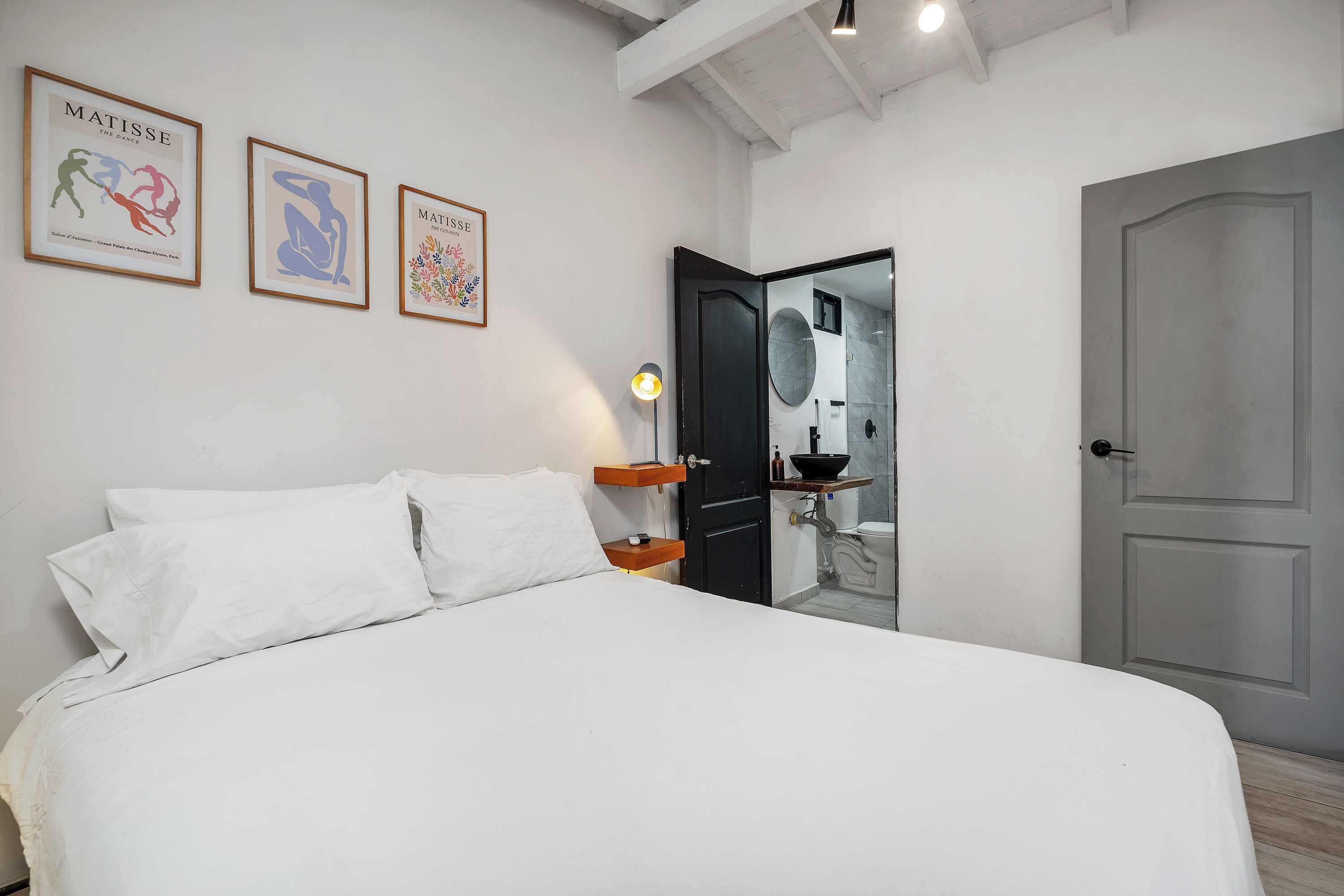 Un dormitorio con cama, almohadas blancas, una mesita de noche, una lámpara y tres cuadros en la pared.