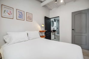Un dormitorio con cama, almohadas blancas, una mesita de noche, una lámpara y tres cuadros en la pared.