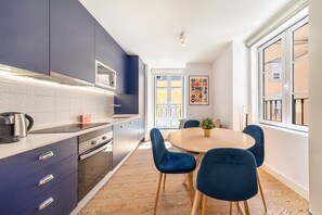 Eine moderne Küche mit einer zentralen Kochinsel, einem Essbereich mit rundem Tisch und blauen Stühlen sowie großen Fenstern mit Ausblick.