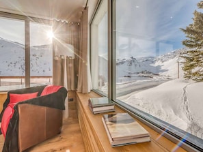Une chambre moderne dotée d’une grande fenêtre donnant sur un paysage montagneux enneigé, d’un canapé en cuir et d’une table en bois sur laquelle sont posés des livres.