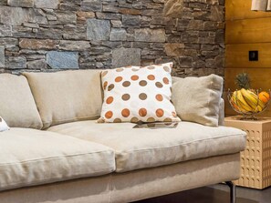 Ein beiges Sofa mit einem Kissen in Punkte-Muster, eine Steinwand und eine Obstschale auf einem Holztisch.