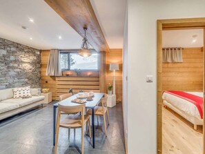 Ein gemütlicher Raum mit Steinwand, Holzbalkendecke, einem Esstisch mit Stühlen, einer Couch und einem Bett mit roter Decke.