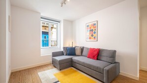 A living room with a grey sofa, a yellow rug, a window with a view, and a colorful abstract painting. - GuestReady - Carolina Building: Ground Floor Left (Lisbon)