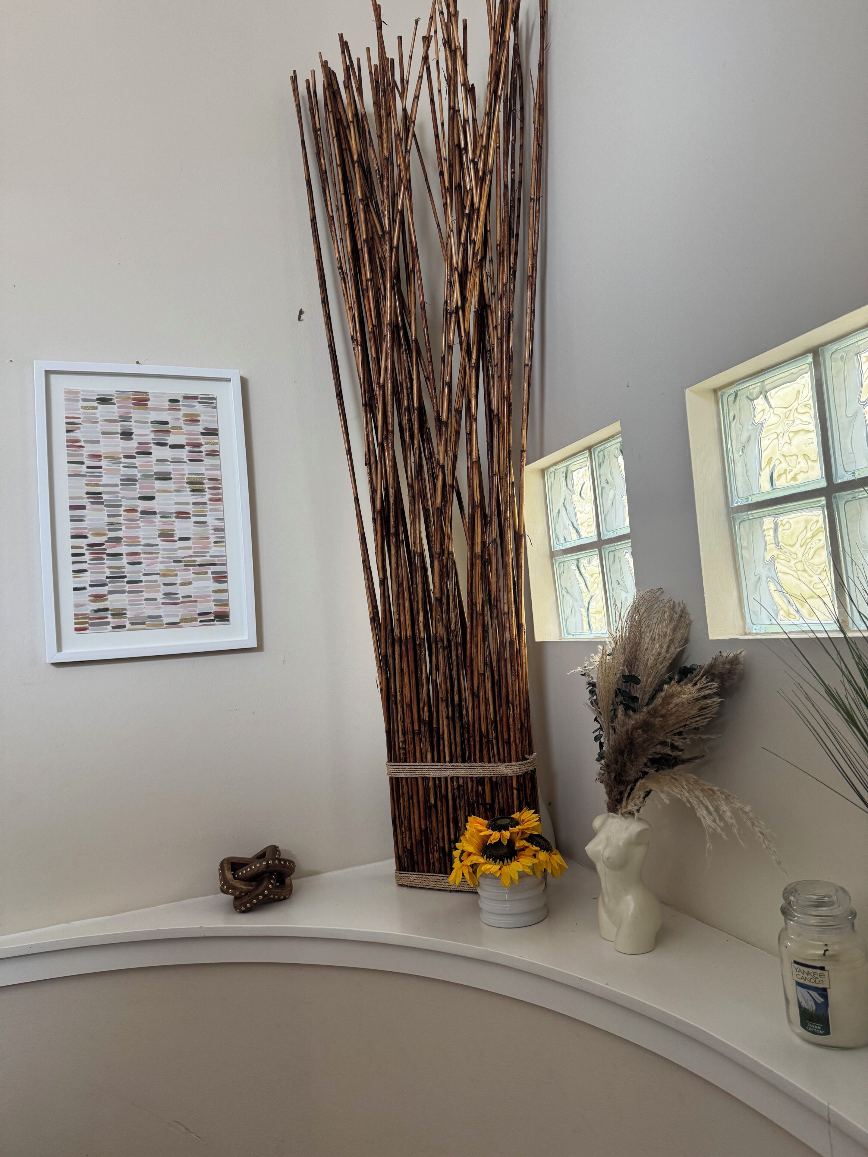 Une pièce avec un mur blanc, une œuvre d’art abstraite encadrée, une haute structure décorative faite de roseaux, un vase avec des fleurs jaunes, une bougie et une fenêtre donnant sur un paysage.