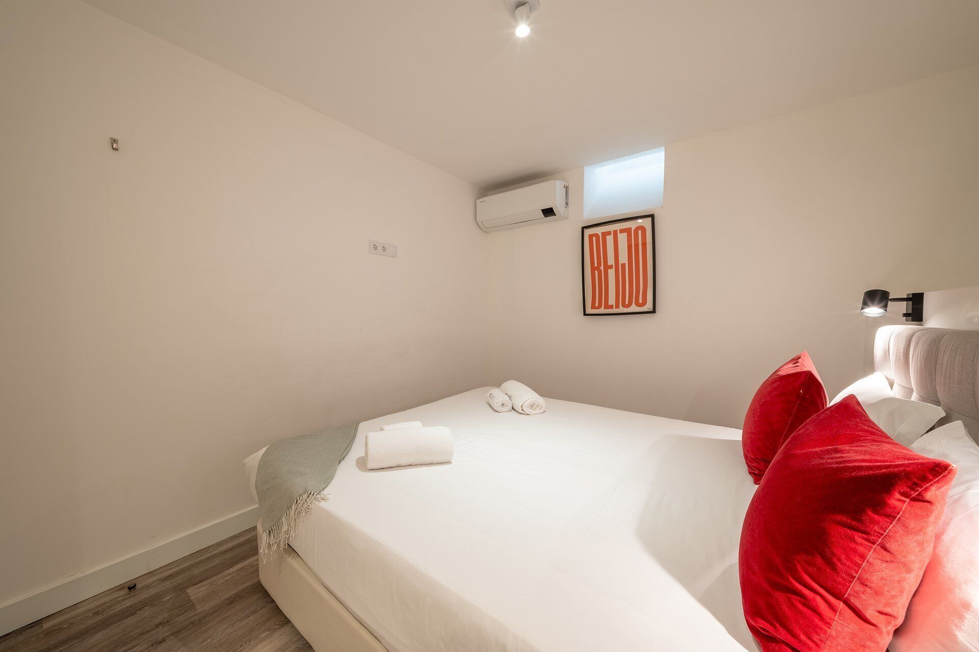 Un dormitorio con cama, aire acondicionado de pared, una obra de arte enmarcada y una lámpara de noche.