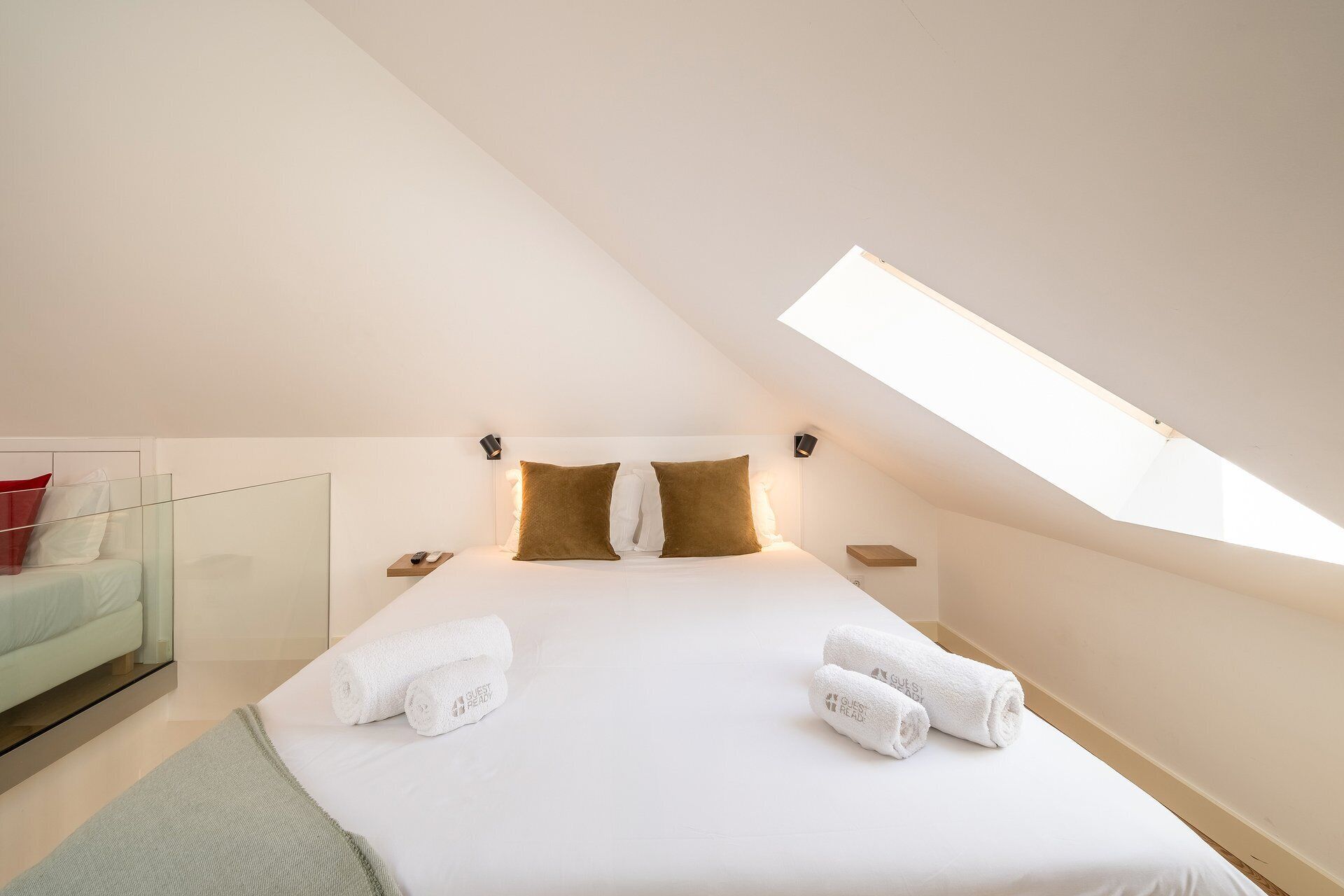 Un dormitorio con tragaluz, una cama con ropa blanca, dos almohadas, una mesita de noche y una pequeña tocador.