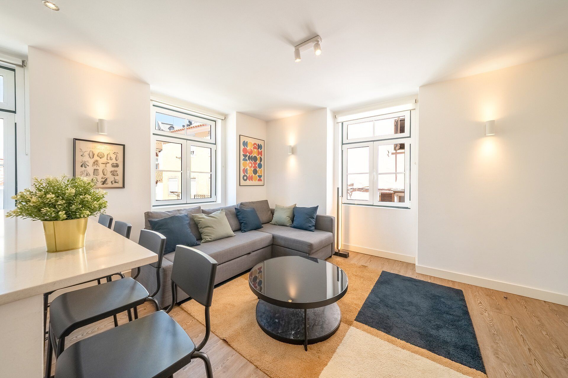 Ein modernes Wohnzimmer mit einem grauen Ecksofa, einem runden Couchtisch und einem Essbereich mit weißem Tisch und schwarzen Stühlen.
