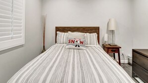 Ein Schlafzimmer mit einem Bett, einem Nachttisch, einer Lampe, einem Fenster mit Jalousien und einer Wandlampe.
