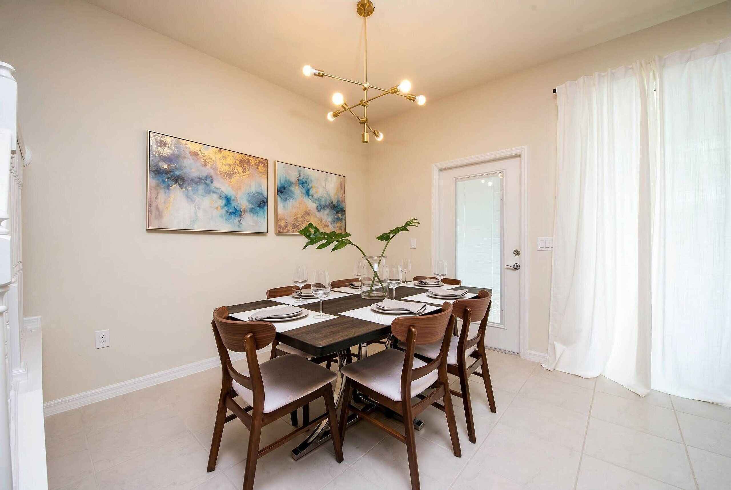 A dining area with a round table, chairs, a chandelier, and two abstract paintings on the wall.