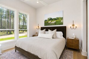 A bedroom with a large bed, a nightstand, a bedside table, a wall-mounted lamp, and a window with a view of trees and grass.