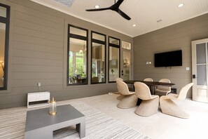 A modern living room with a grey wall, large windows, a ceiling fan, a TV, and a dining area with wicker chairs.