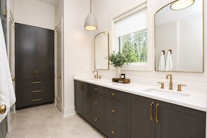 A modern bathroom with dark cabinets, gold handles, a white countertop, and a large mirror.