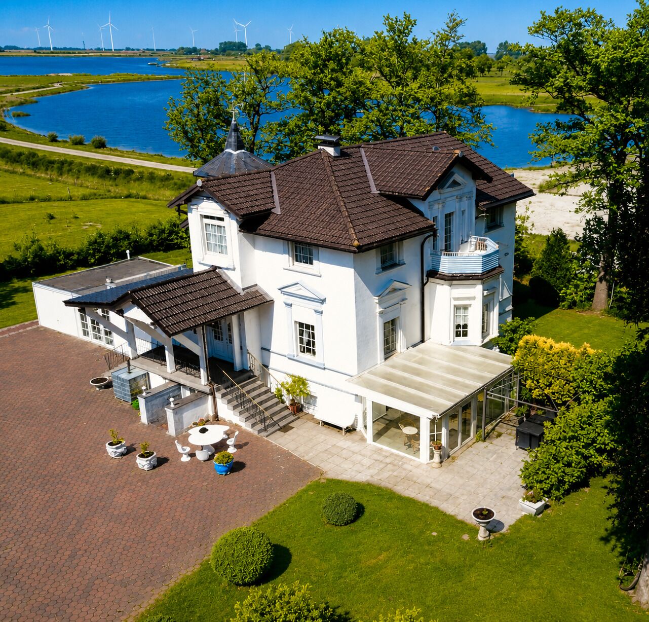 A large white building with a covered porch and a swimming pool, surrounded by lush greenery and a lake with wind turbines in the background.