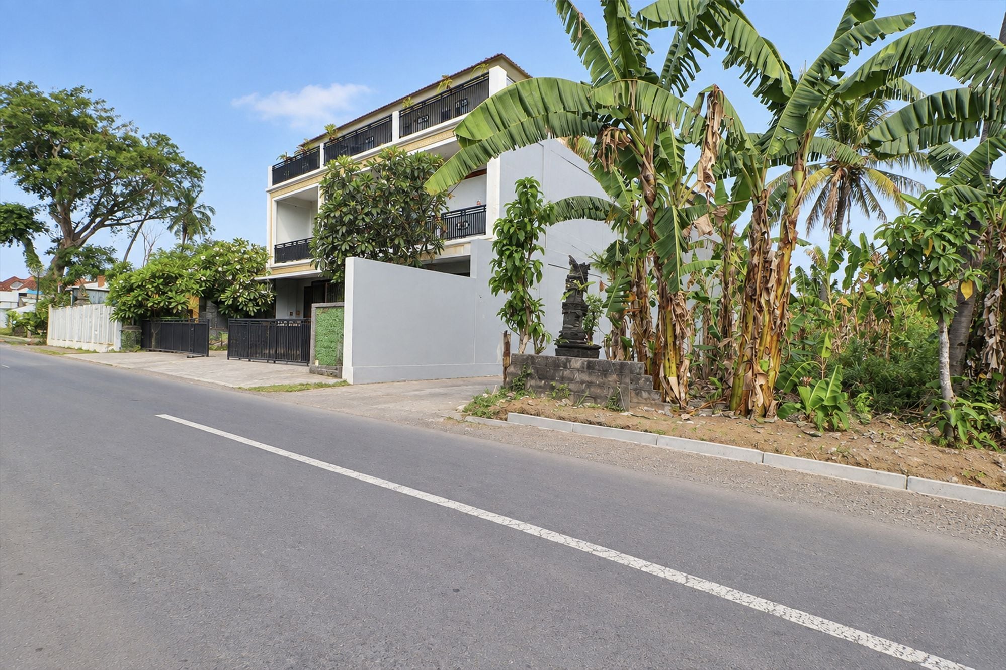 A two-story residential building with a white facade, a balcony, and a garden with banana trees.