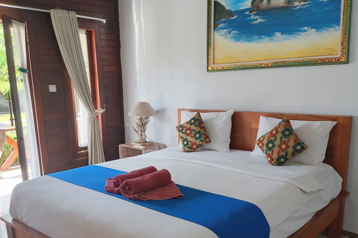 Una cama de madera con ropa de cama blanca y azul, un cuadro de una escena costera y una lámpara sobre una mesita de noche.