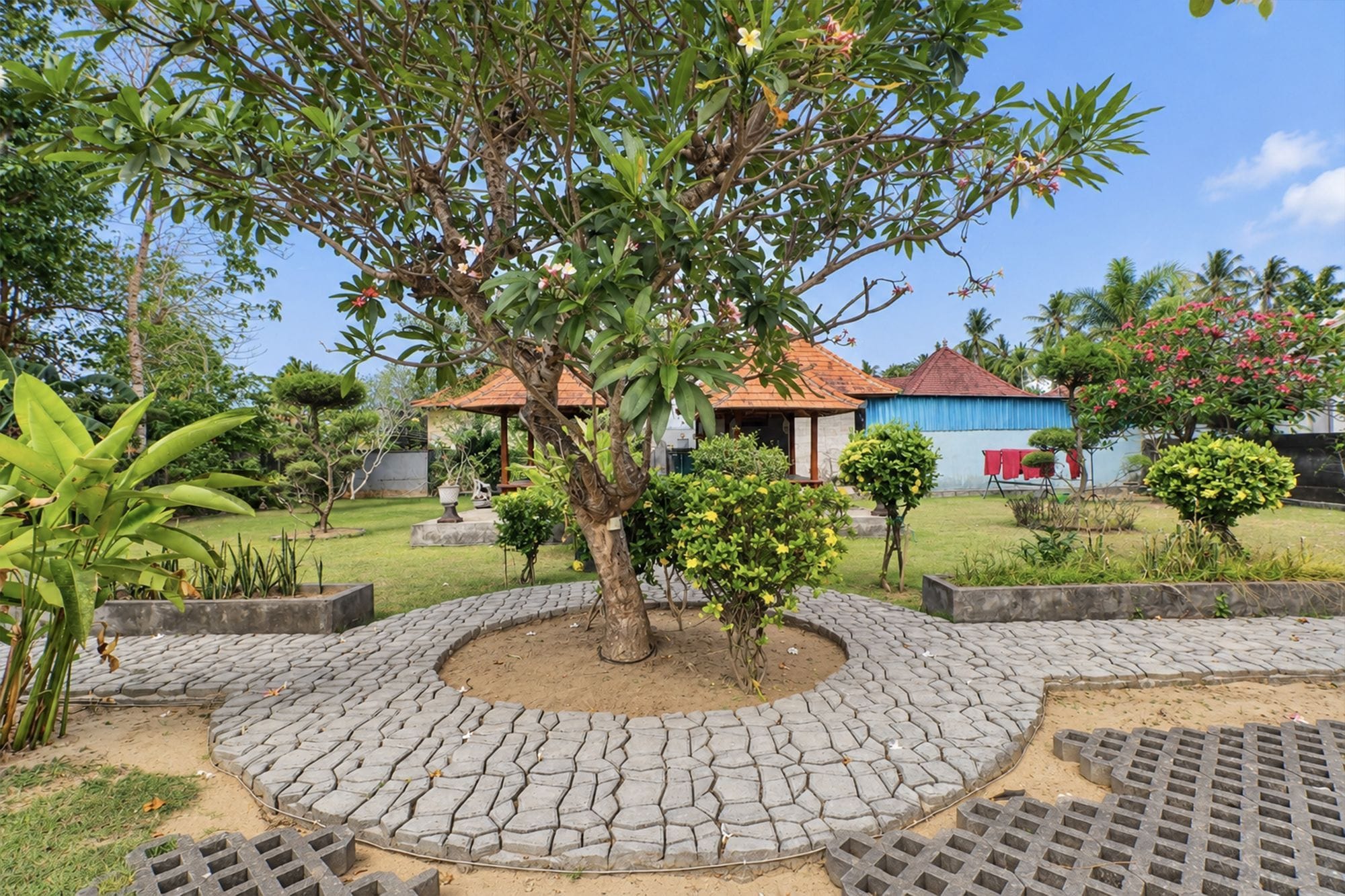 Un jardín con una zona circular empedrada, un árbol y un edificio de techo rojo.