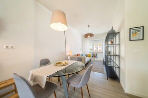 Ein Essbereich mit einem Glastisch, grauen Stühlen, einer beigen Tischdecke, einer Hängelampe und einem Bücherregal mit einem gerahmten Bild.