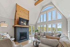 A spacious living room with a fireplace, large windows, and a high ceiling.